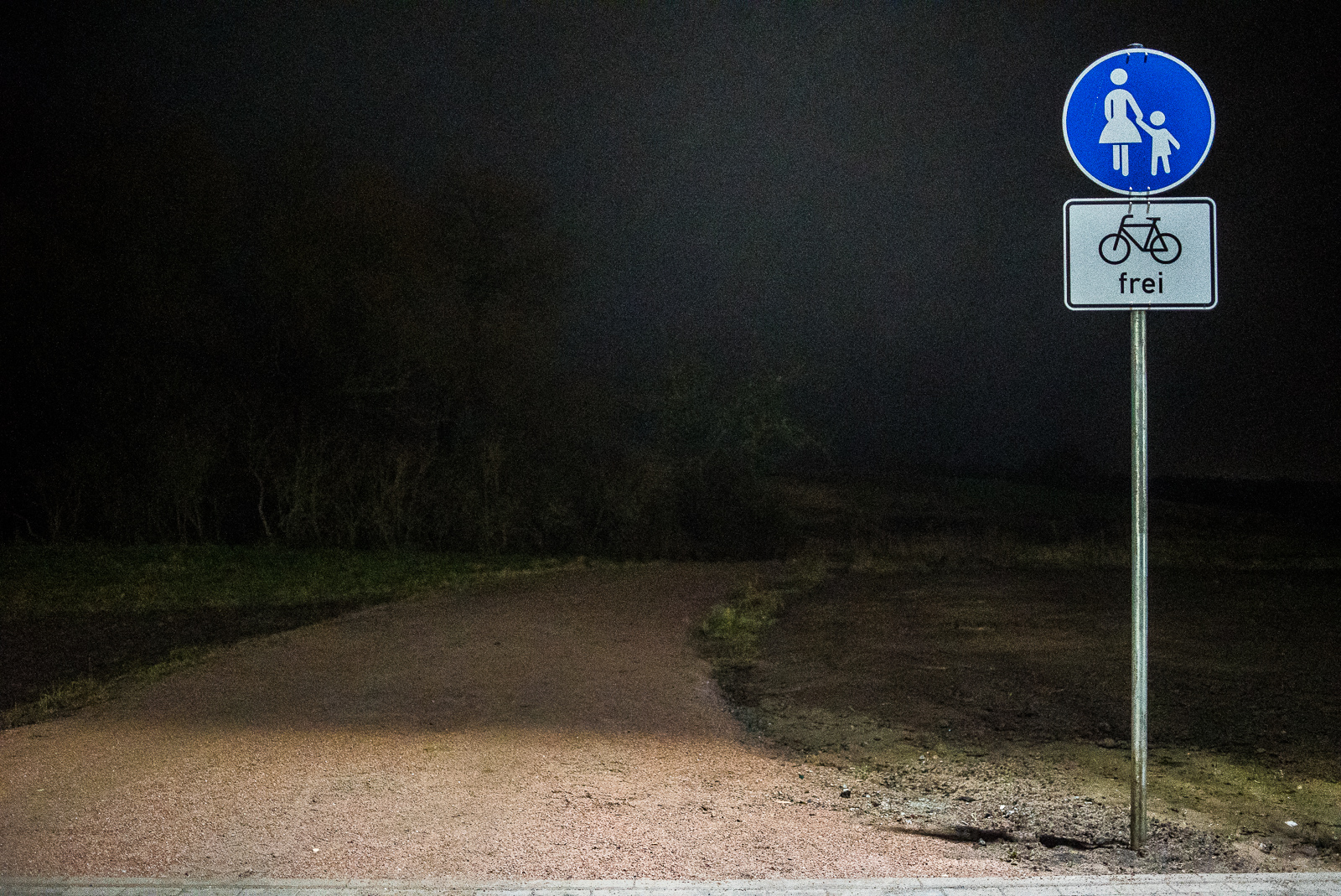 Nachtaufnahme: Fuß-/Radwegschild vor dunkler Wiese