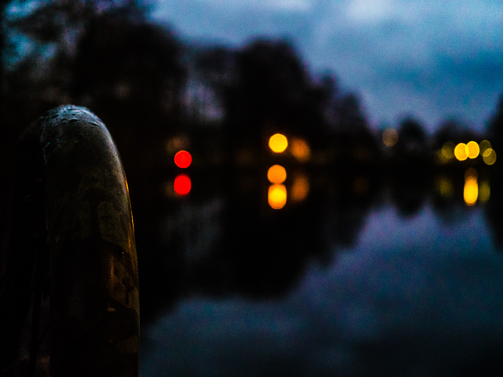 Ansicht eines Teichs im Dämmerlicht, unscharf, nur der metallene Handlauf einer Badetreppe links ist scharf. Eine Baumreihe und darunter diverse Lichter, gelbe und rote, spiegeln sich im Wasser.