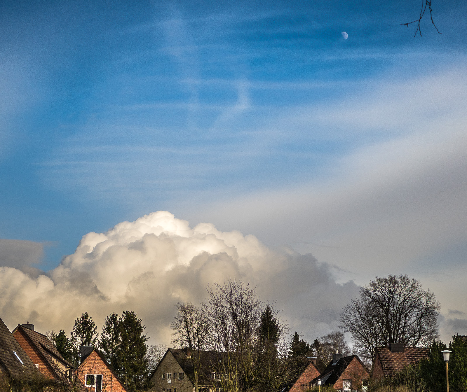 Mächtige sonnenbeschienene Wolke unter blauem Himmel, über kleinen Siedlungshäusern. Oben rechts im Bild der halbe zunehmende Mond, daneben ragt ein Ast ins Bild.