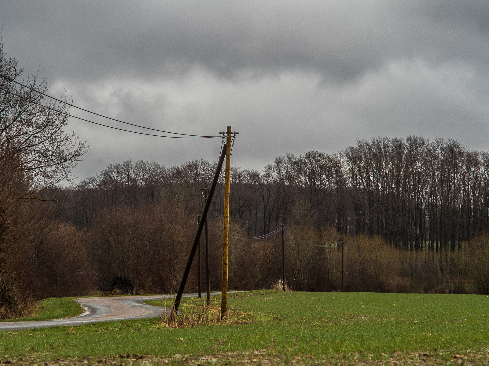 Blick in die Tiefe einer Landschaft längs einer von links ins Bild laufenden Landstraße, die von einer einfachen Leitung mit zwei Kabeln und dünnen Pfosten begleitet wird.