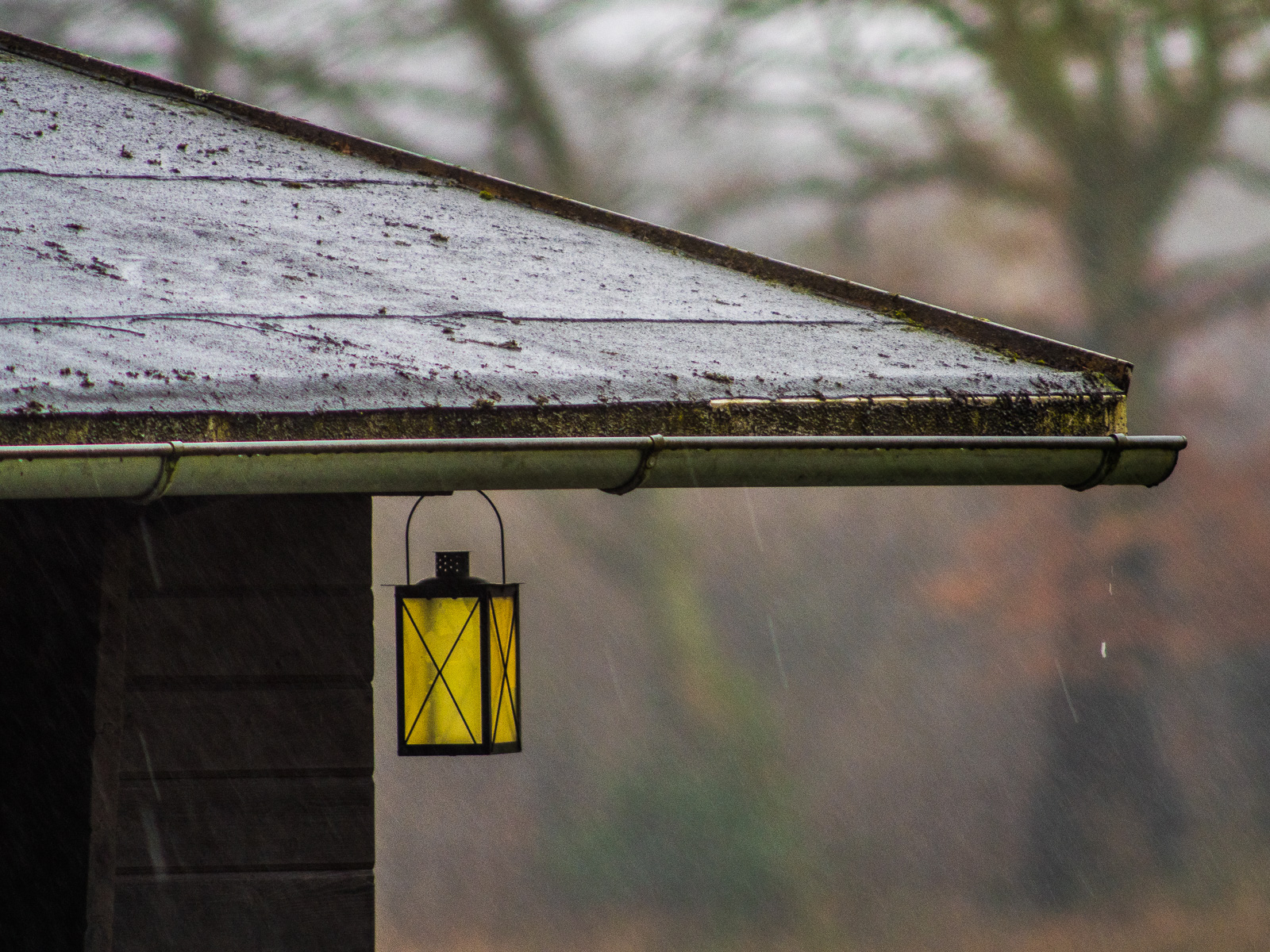 Das Blechdach einer einfachen Hütte im Regen. Unter dem Dachvorsprung hängt eine Laterne mit leuchtend gleber Verglasung.