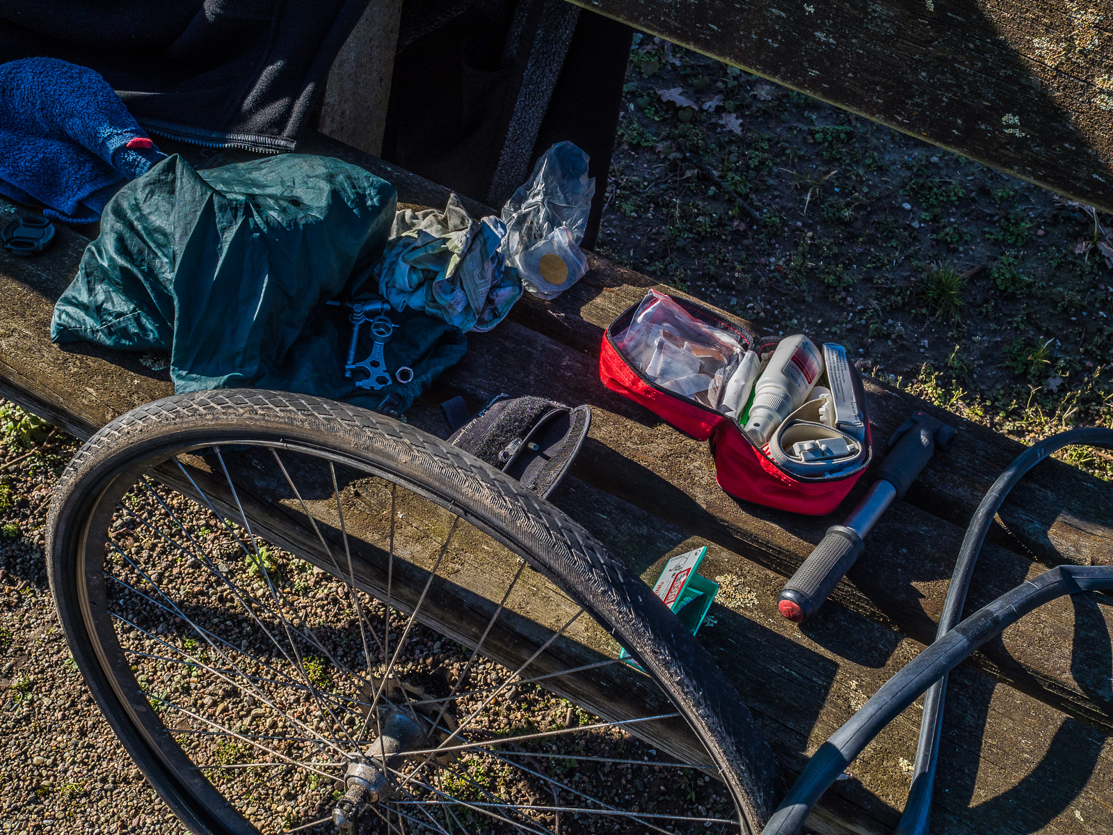 Ein ausgebautes Fahrrad-Hinterrad lehnt an einer Parkbank,drauf ausgebreitet diverse Werkstatt-Utensilien, eine Luftpumpe, ein Fahrradschlauch und ein kleiner Erste-Hilfe-Beutel.