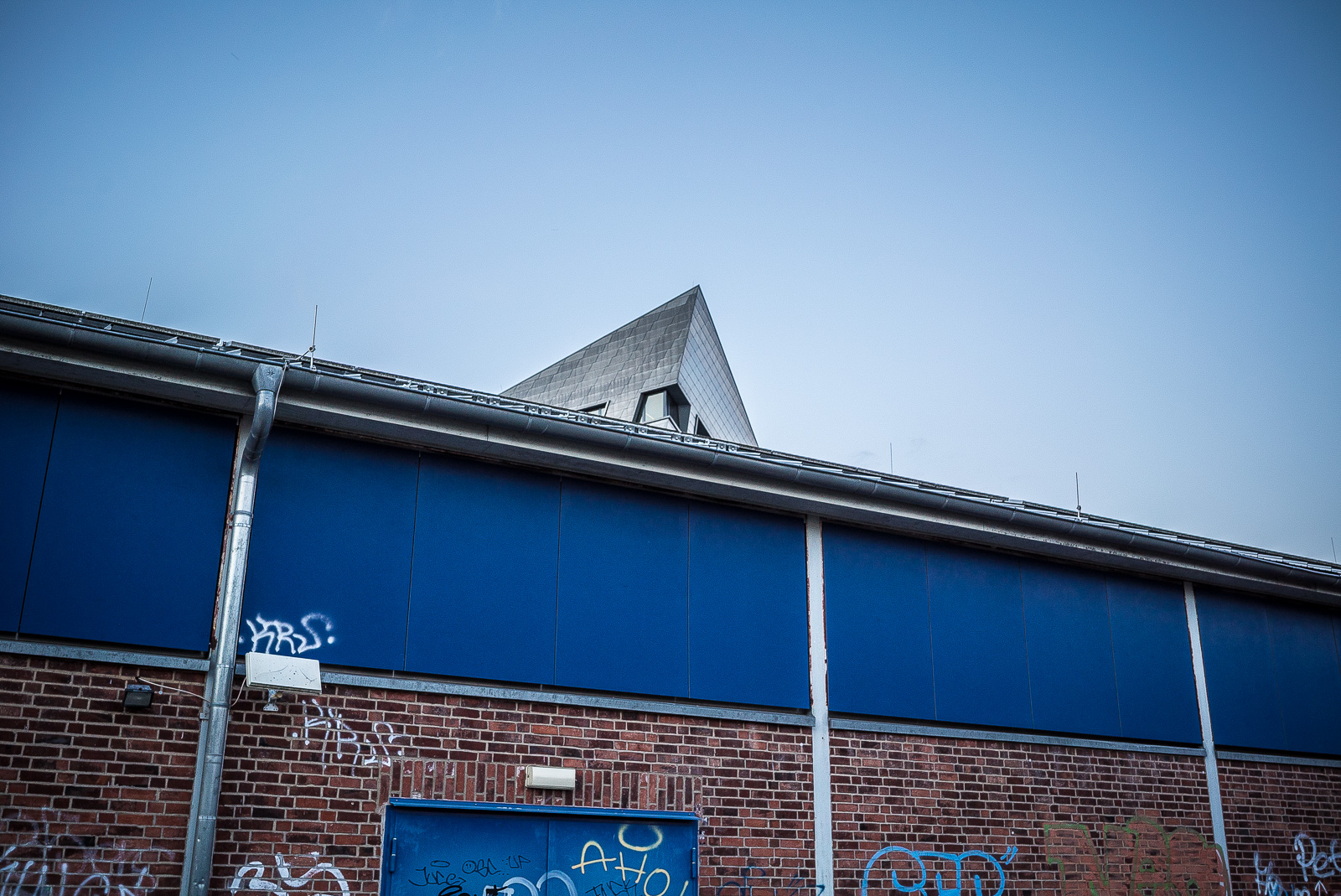 Die Spitze des Hauptgebäudes der Leuphana Universität Lüneburg schaut über dem Dach eines unten geklinkerten, oben blau verkleideten, mit Graffiti beschmierten Schuppens hervor