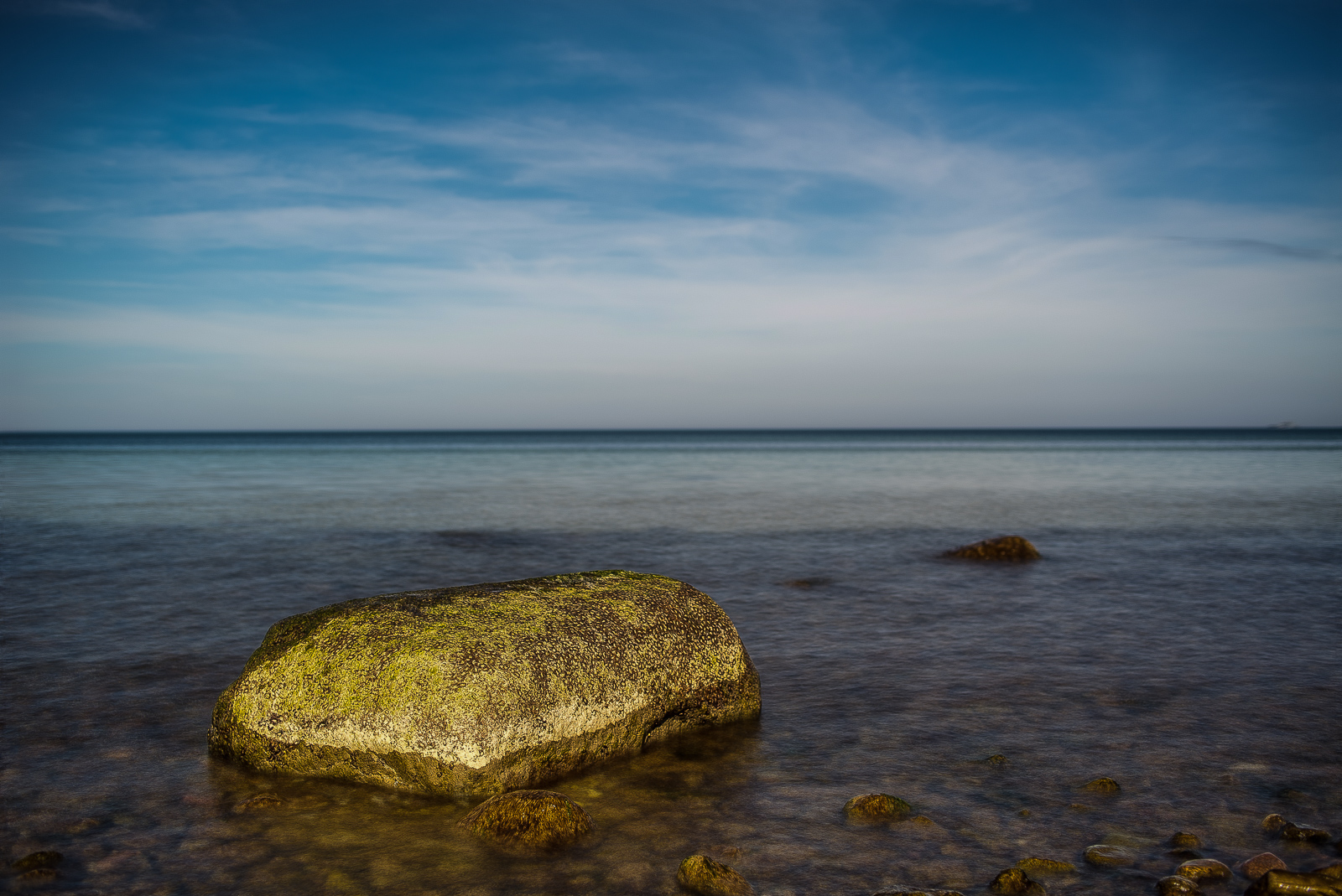 Meerlandschaft. In der Bildmitte der Horizont, vorn links ein großer (1/3 Bildbreite) von der Sonne angestrahlter Felsen nahe des Ufers