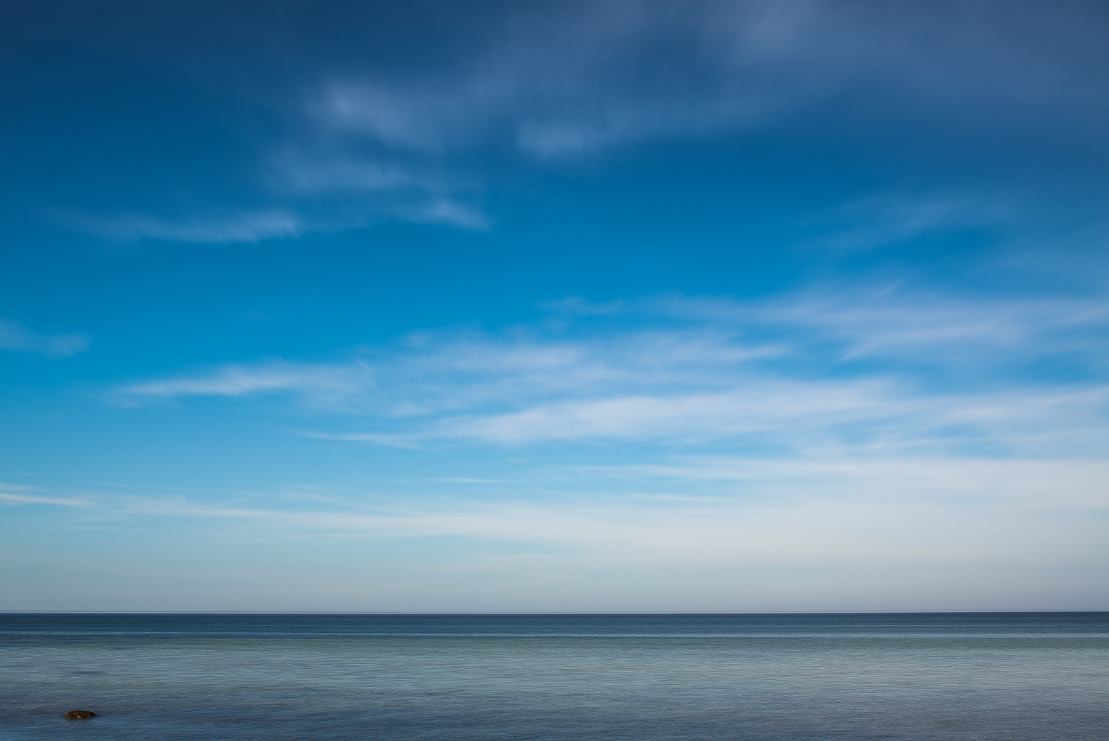 Das Meer unter tief blauem Wolkenhimmel. Der Horizont liegt sehr nah dem unteren Bildrand, ganz links unten unterbricht ein einzelner kleiner Felsen die ansonsten glatte Wasseroberfläche