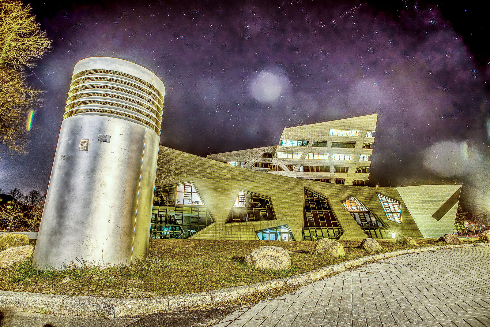 Universität Lüneburg, Hauptgebäude: Der futuristische Bau in kontrastreicher Bearbeitung unter Sternenhimmel. Im Vordergrund ein metallisches Gebilde, vermutlich Belüftung einer Tiefgarage