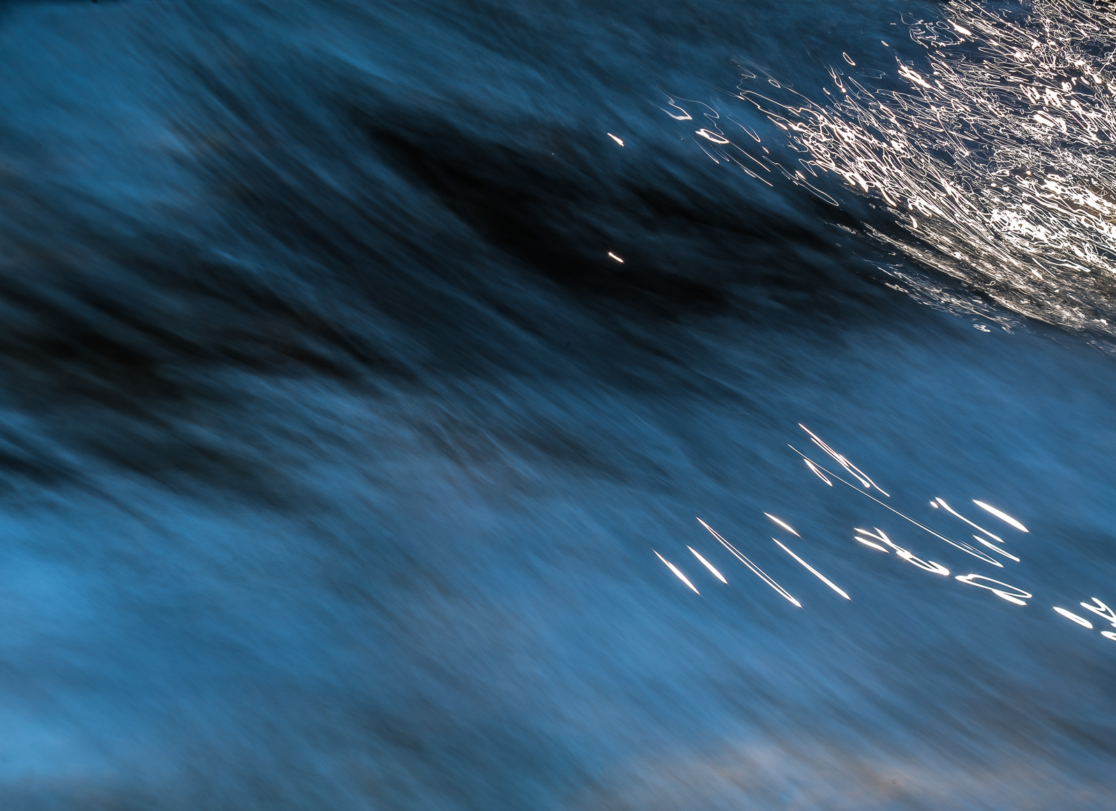 Eine diffuse blaue Wasseroberfläche, lange belichtet, die Bewegungsrichtung des Wassers von links oben nach rechts unten. Rechts oben und mittig mehrere krakelig aussehende Lichtreflexe, wie mit dem Pinsel und Deckweiß gezeichnet.
