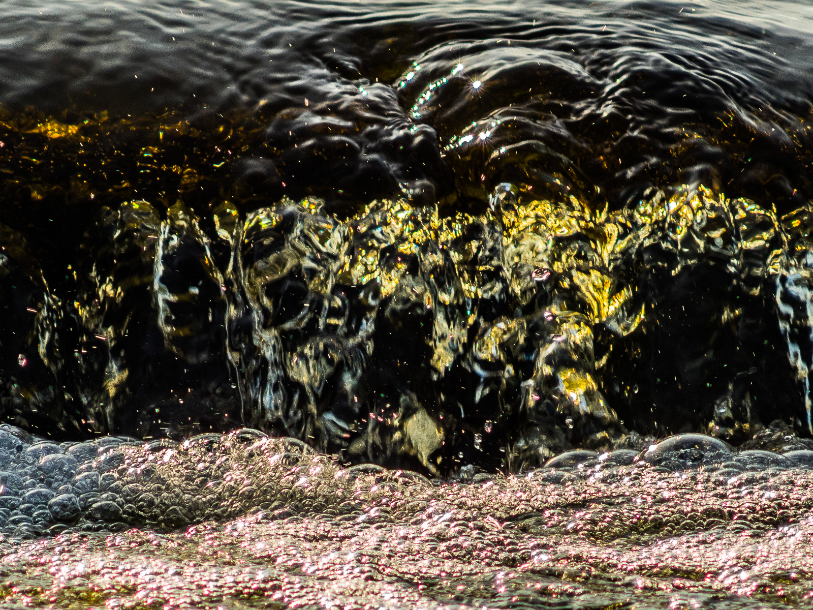 An einer kleinen Staustufe sprudelt das dunkle Wasser eines Moor-Bachs herab, unten türmen sich Blasen auf.