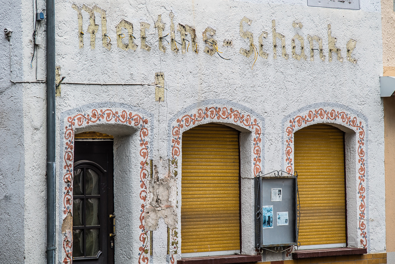 Fassade einer geschlossenen Kneipe, zwei Rollläden, man erkennt die Spuren der vormaligen Beschriftung Martins-Schänke