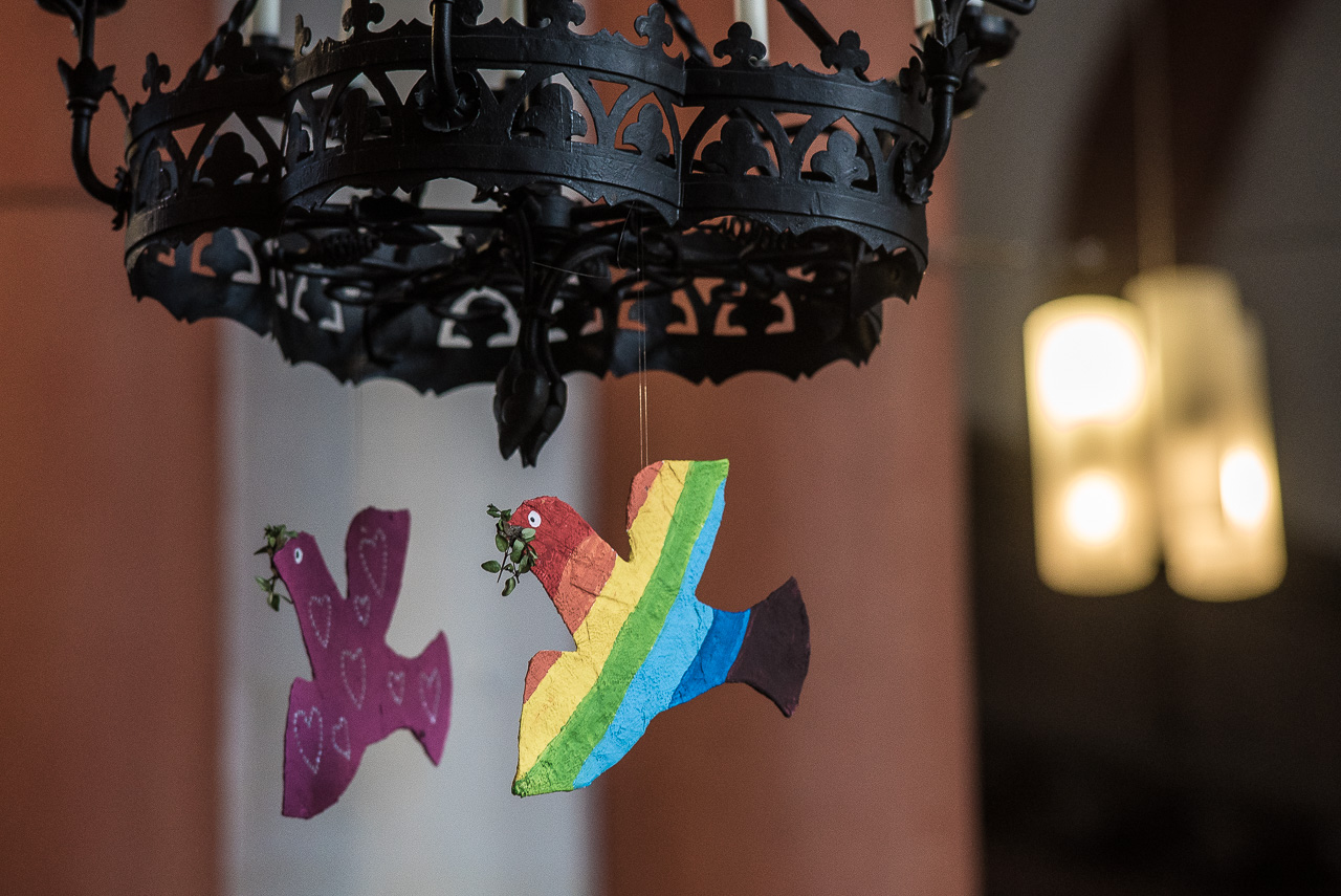 An einem runden eisernen Kerzenhalter in der Kirche hängen zwei bemalte Friedenstauben, eine lila mit glitzernden Herzchen, eine in Regenbogenfarben.