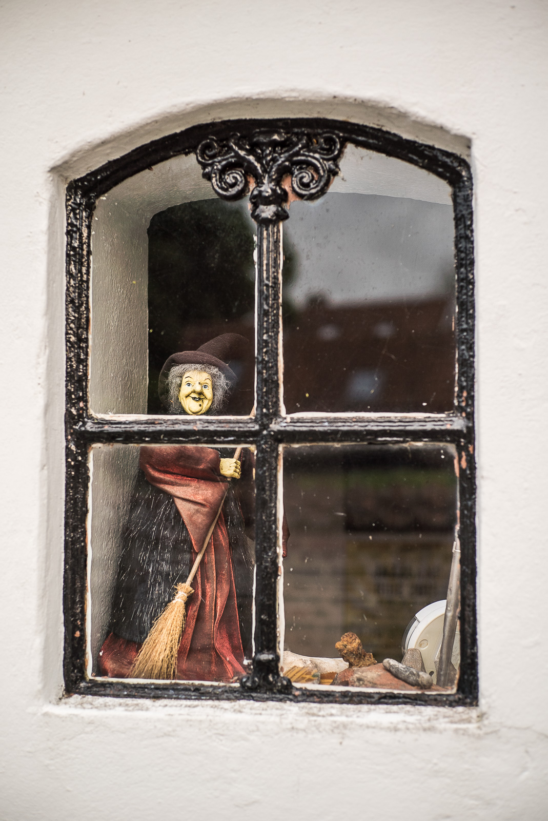 Durch ein metallgefasstes Fenster schaut uns die rot gekleidete Figur einer Hexe mit Besen an