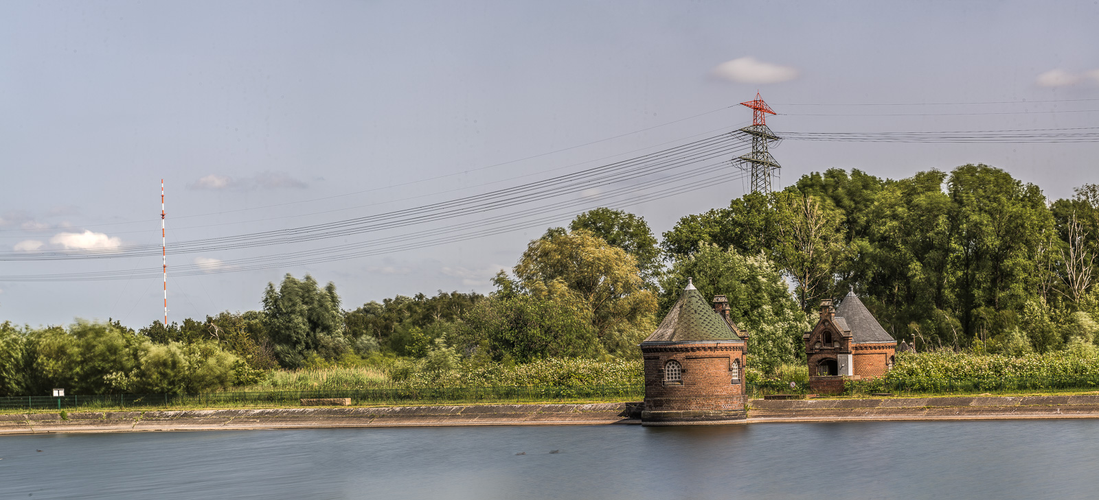Wasserkunst Kaltehofe Hamburg: Vorn ein Wasserbecken, zwei Häuschen, dahinter Wald, am Horizont ein Strommast und ein weiß-rotes Funkfeuer