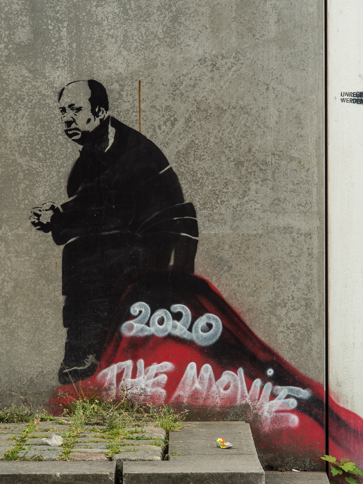 Eine graue Betonmauer. Eine gesprayte Figur eines schwarz gekleideten, traurig wirkenden Mannes , undefinierter roter Untergrund. Weiße Krakelschrift: "2020 The Movie"