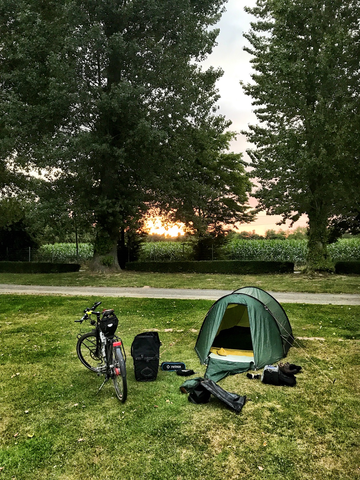 Rasenfläche eines Zeltplatzes, hinter Bäumen geht die Sonne auf. Ein grünes Tunnelzelt, ein Fahrrad und mehrere Packtaschen.