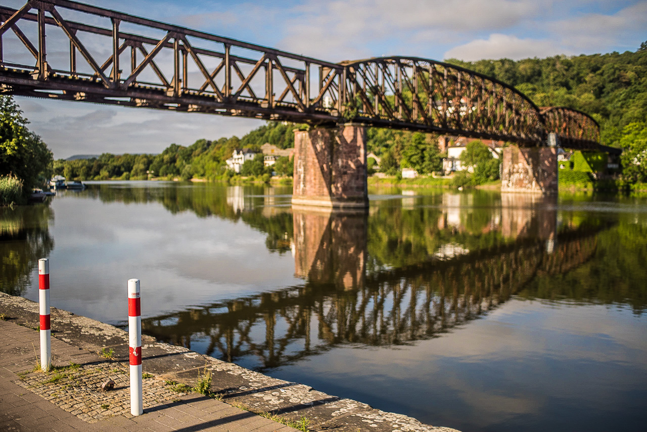 eine Stahlgitter-Eisenbahnbrücke über einen Fluss, im Vordergrund zwei weiß-rote Poller