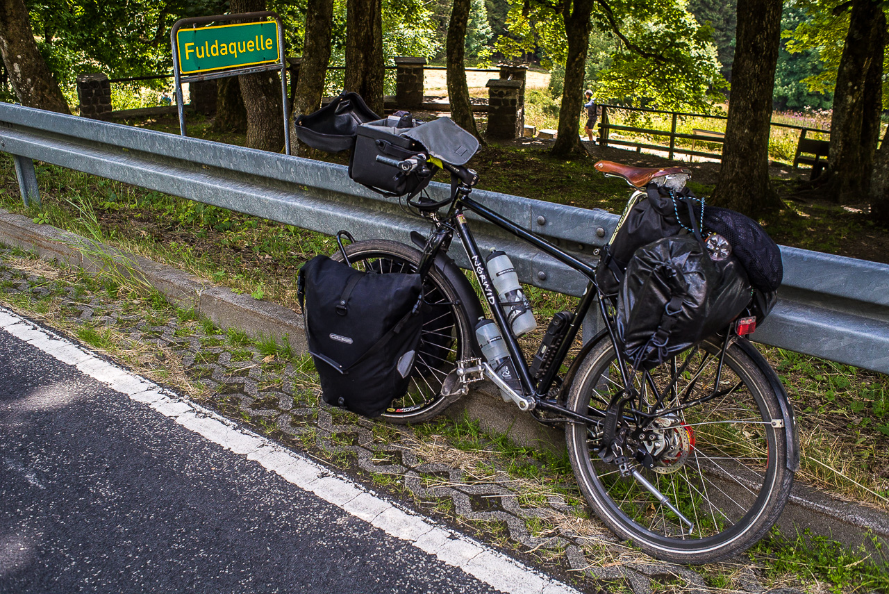 Ein Fahrrad lehnt an einer Straßenbegrenzung, dahinter ein Schild "Fuldaquelle"