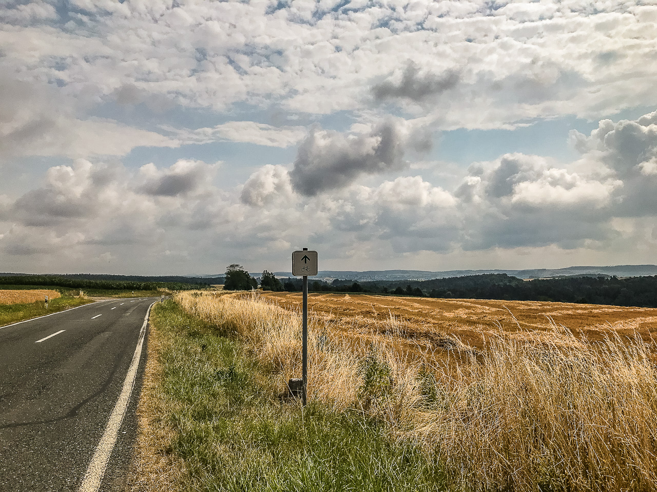 Eine Landstraße führt durch abgeerntete Felder unter kräftigen Wolken bergab