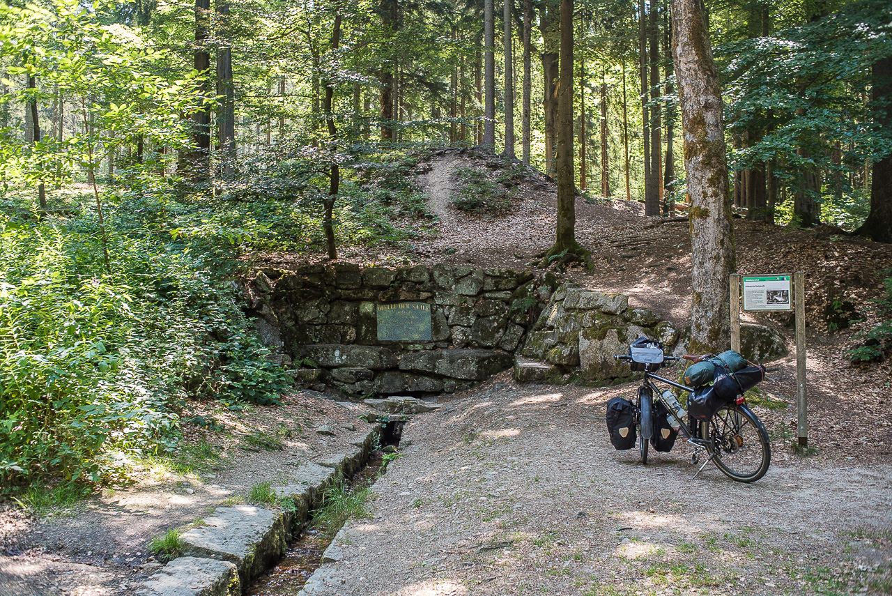 Ein Fahrrad vor einer steingefassten Quelle in einem Bergwald