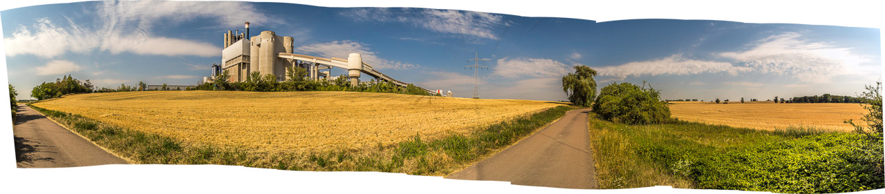 Panorama einer Landstraße, die durch Getreidefelder an einem Zementwerk vorbeiführt