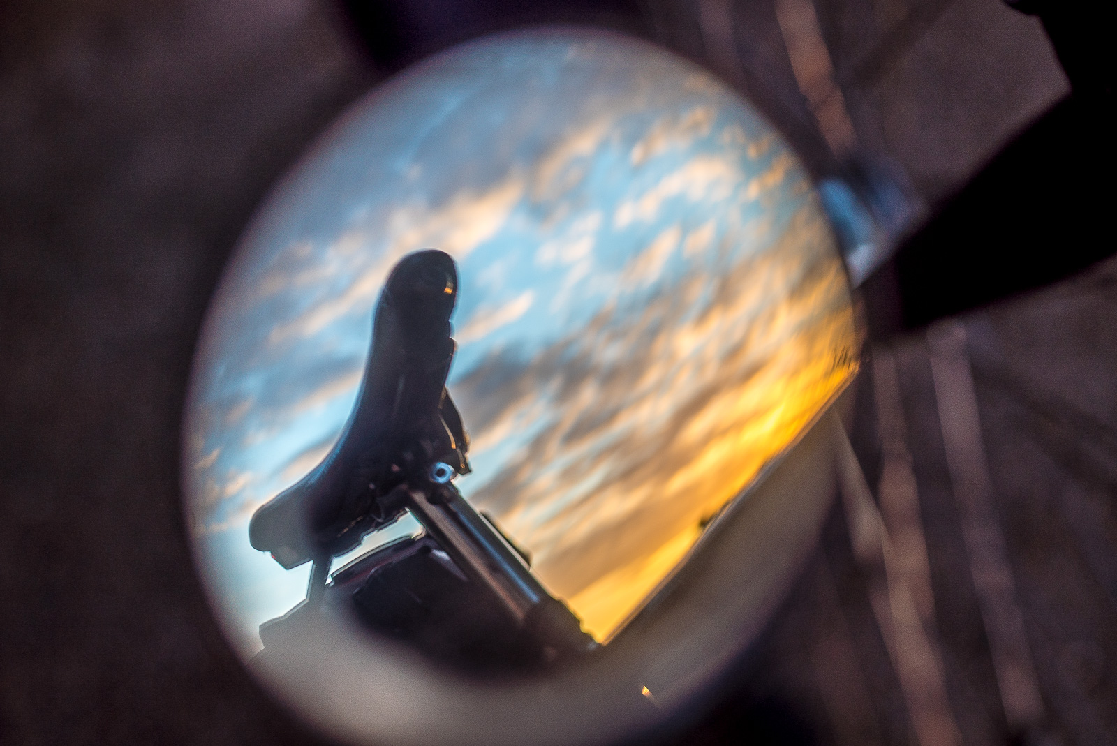 ein FahrradSattel in einem runden RückSpiegel, dahinter leuchtende Wolken bei SonnenUntergang