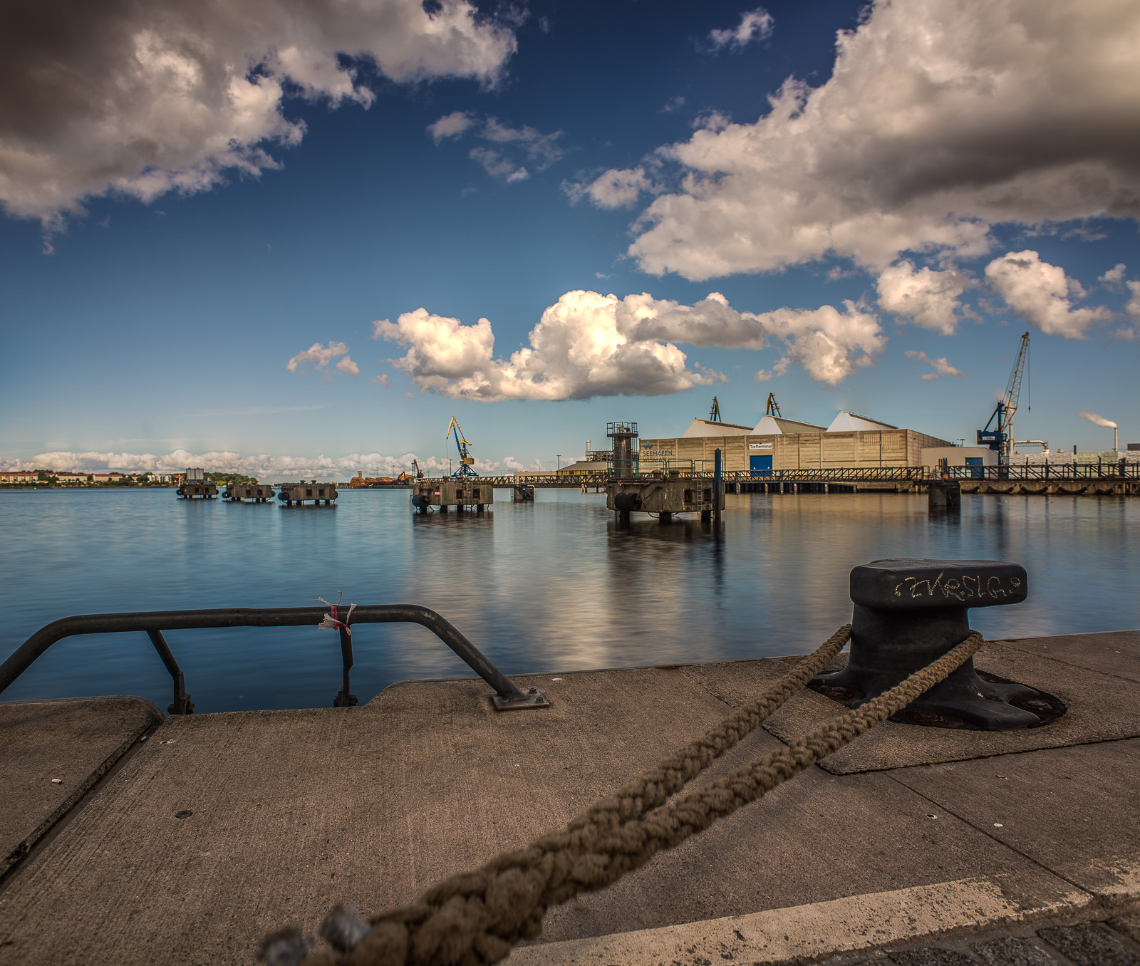 Weitwinkelfoto eines Pollers am Hafenbecken. Im Hintergrund andere Anlagen zum Festmachen von Schiffen und ein Lagerhaus mit Kränen. Darüber viele Wolken.