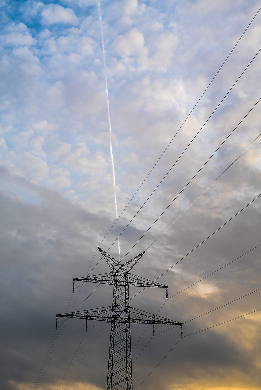 Ein StromMast vor HäufchenWolken. Ein KondensStreifen verläuft so über den Himmel, dass er den StromMast mittig zu treffen scheint.