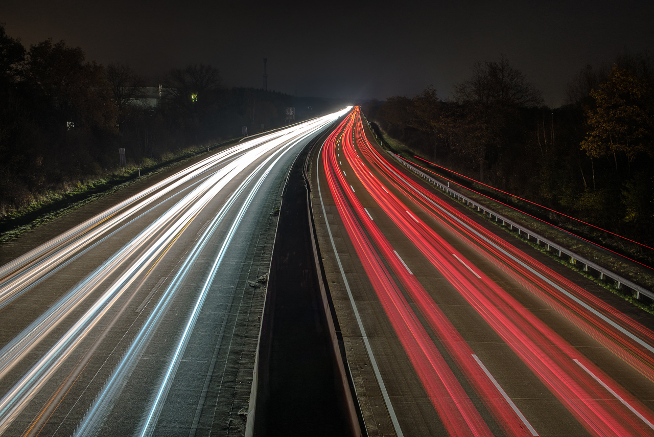 NachtAufnahme einer dreiSpurigen Autobahn von oben mittig – links die weißen bis gelben LichtSpuren entgegenKommender Autos, rechts rote LichtSpuren von RückLichtern