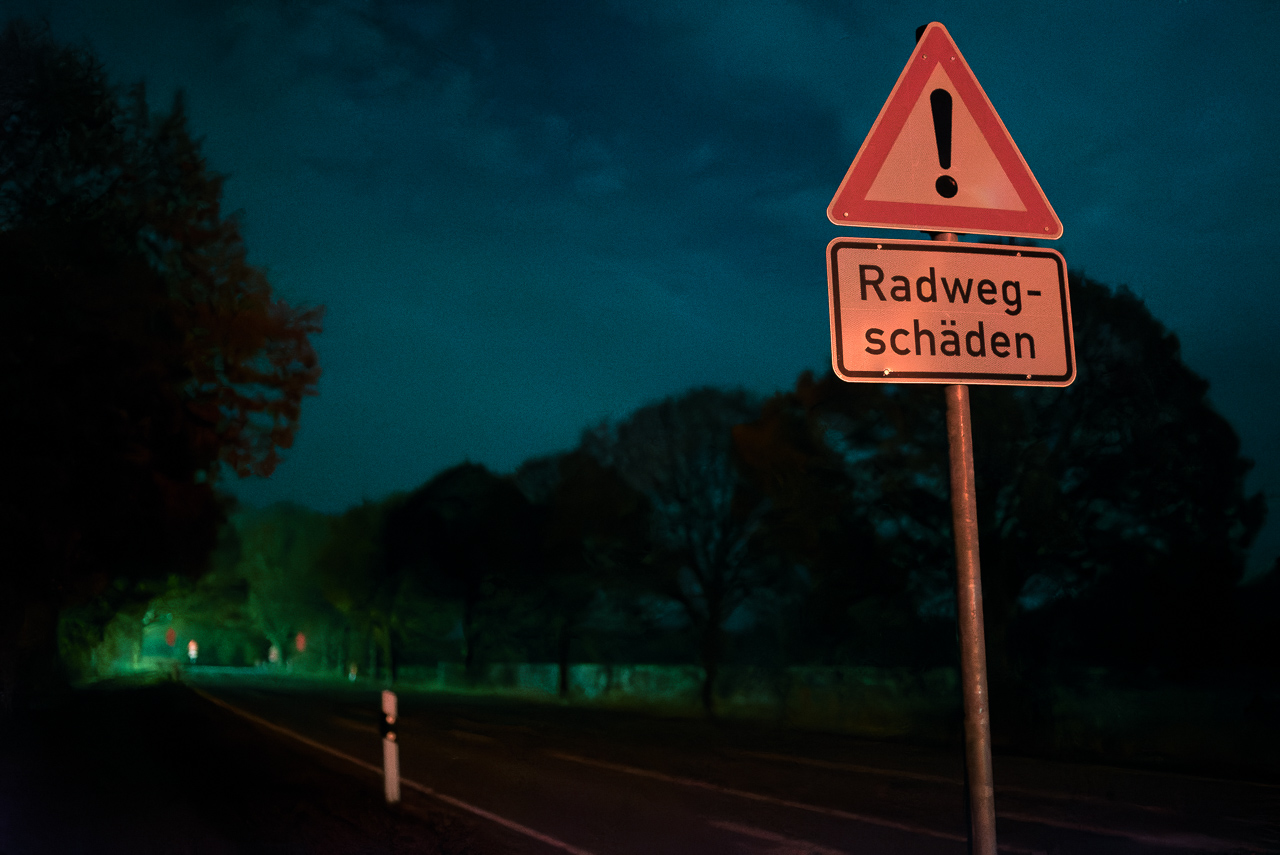 Nachtszene, eine Straße, von Bäumen gesäumt. Ein Schild Achtung! Radweg-Schäden" wird im VorderGrund rot beleuchtet. Sehr dunkel, im HinterGrund künstlich wirkendes grünes Licht.