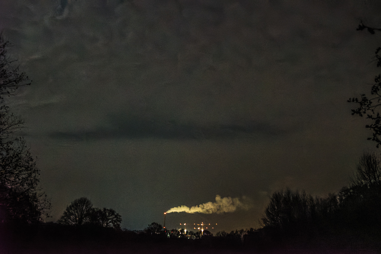 Sehr Dunkler WolkenHimmel, am unteren BildRand Silhouetten von Bäumen, mittig ein rauchender SchornStein und mehrere beleuchtete BauKräne
