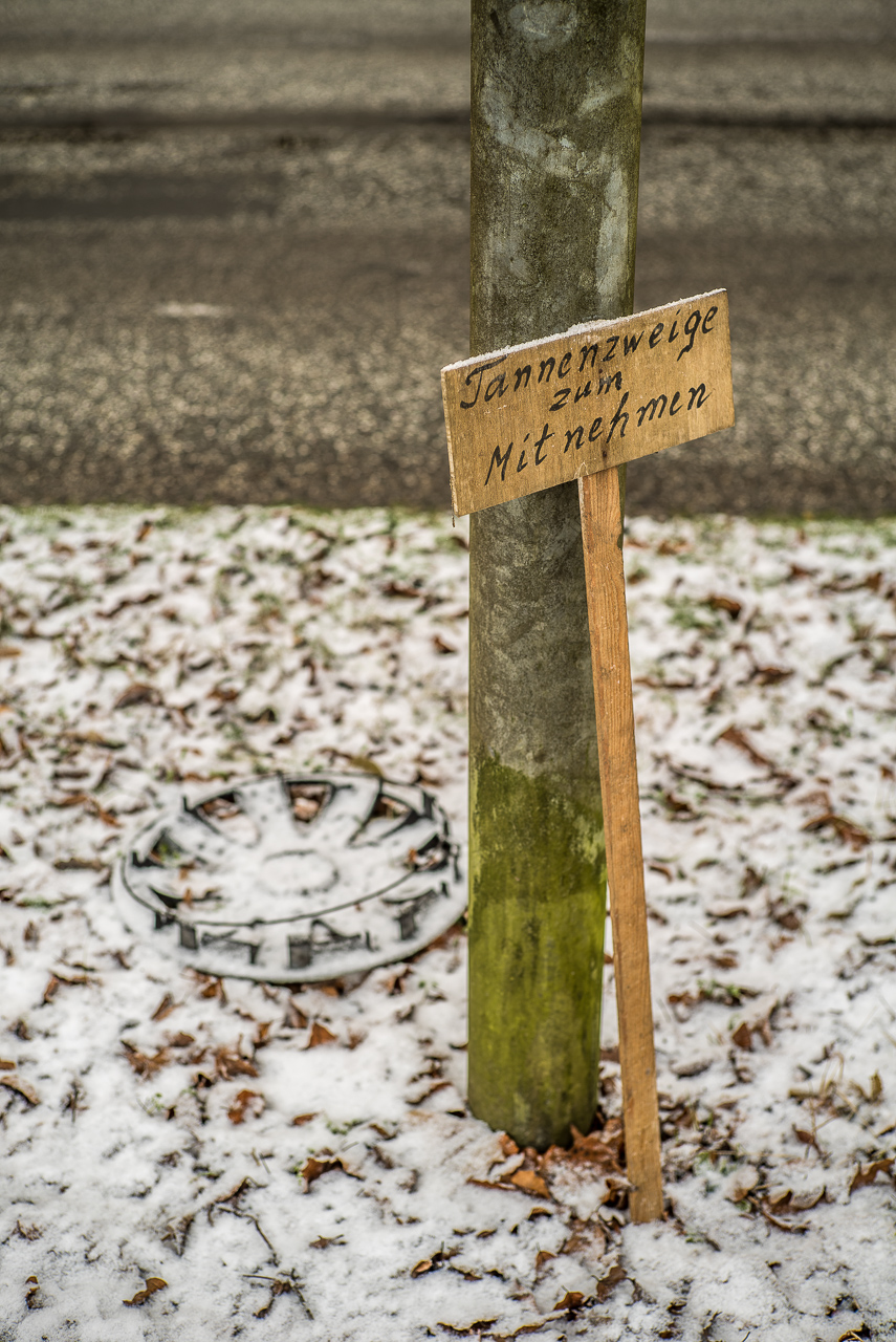 An einem Pfosten im Schnee neben einer Straße lehnt ein grob gezimmertes Schild "Tannenzweige zum Mitnehmen". Dahinter liegen keine Tannenzweige, aber eine verschneite Radkappe