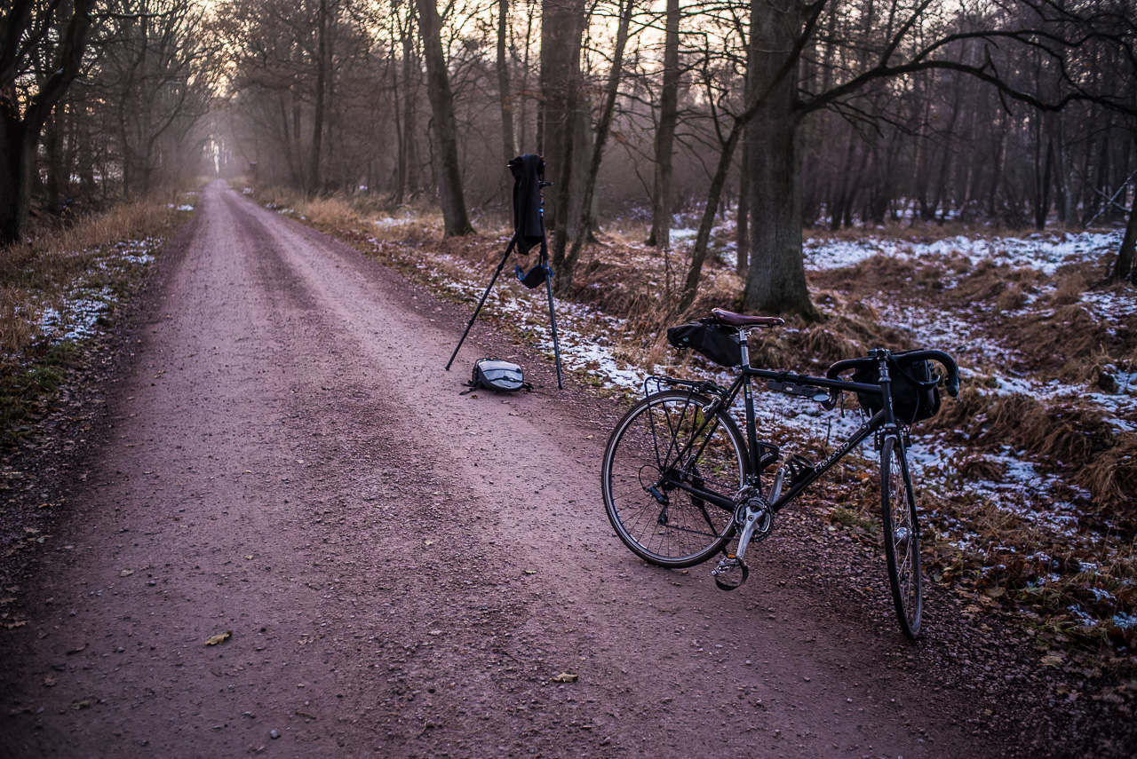 Ein geschotterter Fahrweg führt diagonal durchs Bild. Er ist von Bäumen und Wiesen gesäumt, es liegt etwas Schnee. Am rechten vorderen Rand steht ein schwarzes Fahrrad, dahinter ist ein FotoStativ aufgebaut.