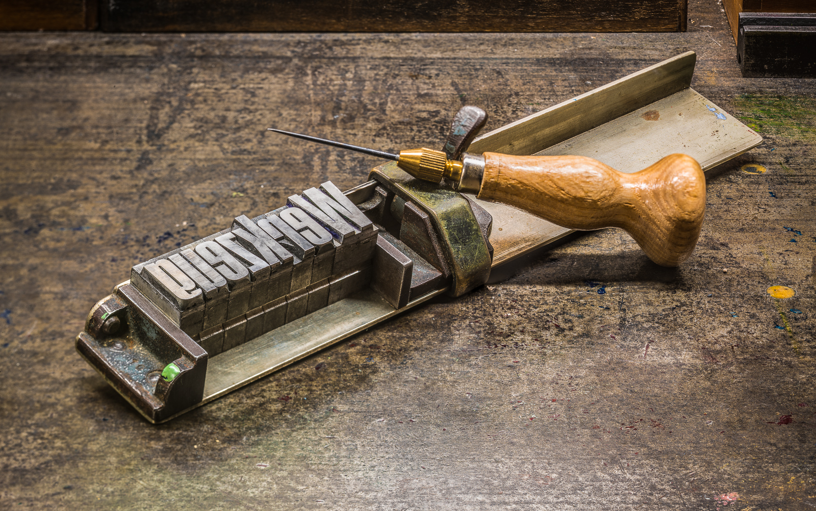 Ein WinkelHaken aus dem Handsatz-Bereich sowie eine Ahle liegen auf einer metallenen WerkBank. Im WinkelHaken steckt aus schmalen, großen BleiLettern das Wort "Werkzeug".