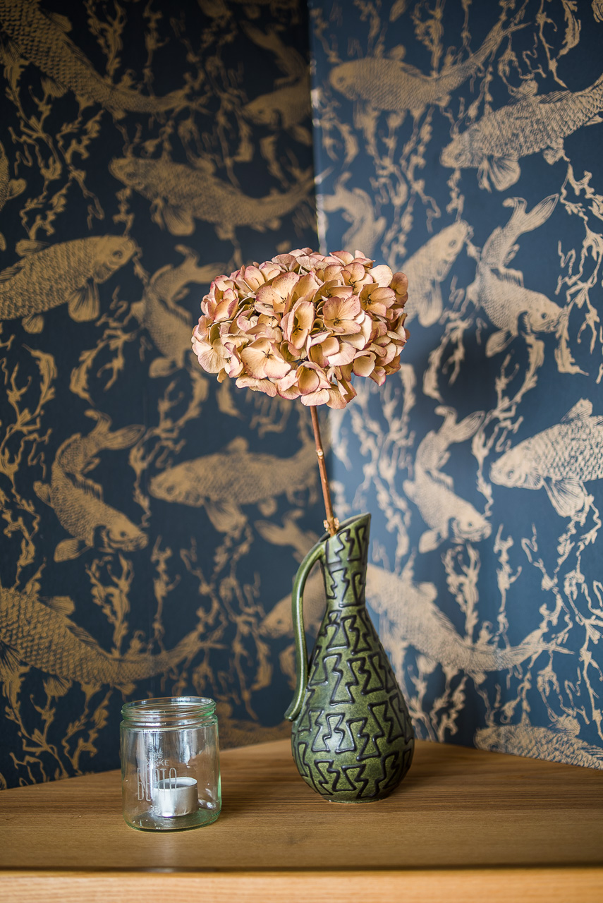Eine getrocknete HortensienBlüte in grüner schlanker Vase vor einer blauen Tapete mit goldenen Fischen.