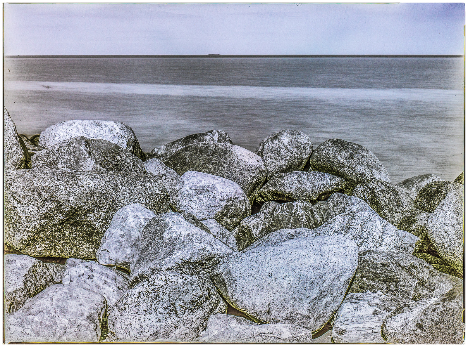 Querformat einer KüstenLandschaft. Der Horizont liegt nah am oberen BildRand. Darunter sanft dünendes Wasser, vorn sehr große runde Steine, die als WellenBrecher am Strand aufgetürmt sind. Das Bild ist schwarz-weiß mit leichter blau/brauner Tonung.