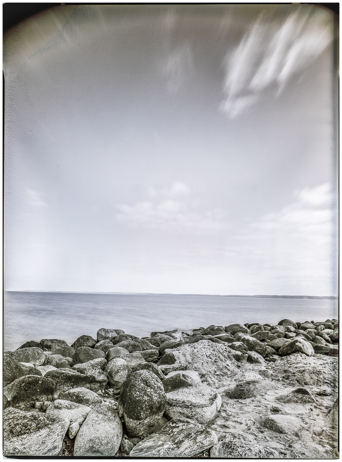 HochformatAufnahme einer Reihe von Steinen am Strand, die sich diagonal von vorn links in die Tiefe des Bildes ziehen. Darüber ein fast klarer Himmel mit wenigen Wolken. Zarte Farbigkeit zwischen HimmelsBlau und SandBraun