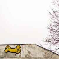 Weiße Hausmauer vor weißem Himmel, auf der Wand ein gelbes Graffito: Ein Arm, der an beiden Seiten je eine geballte Faust hat