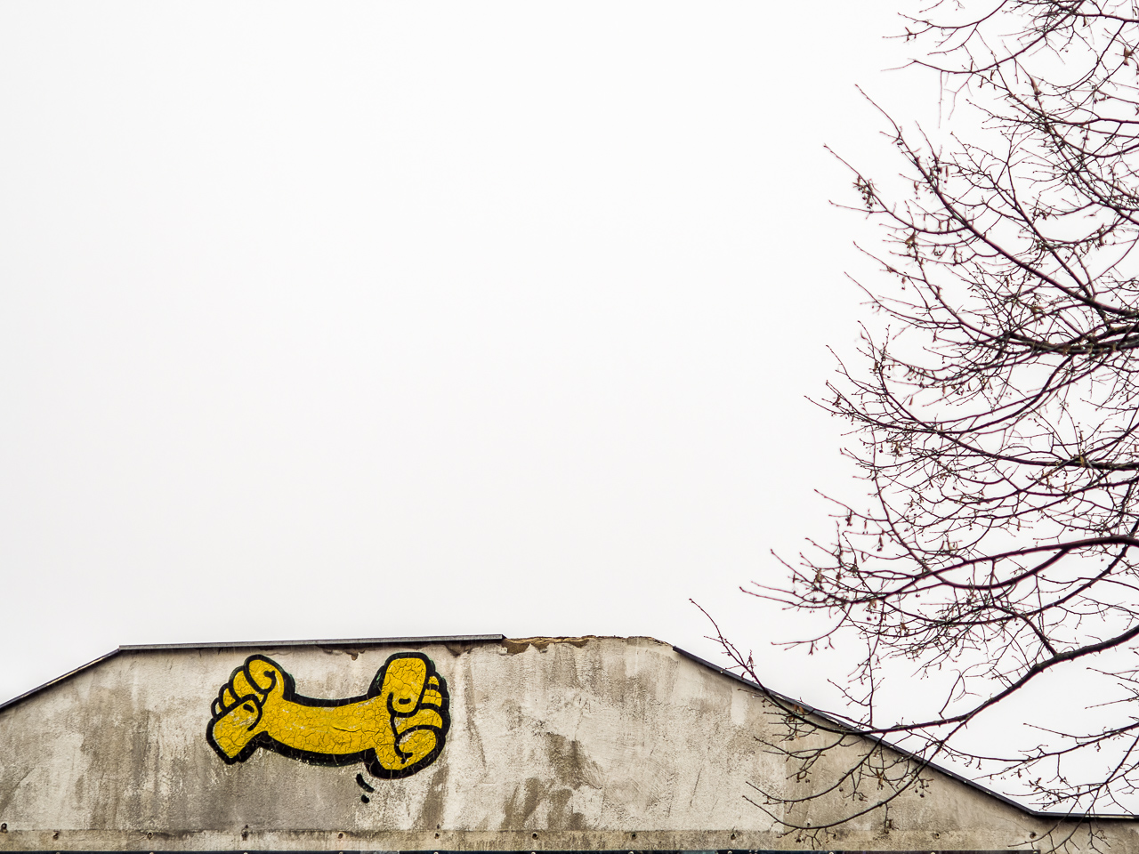 Weiße Hausmauer vor weißem Himmel, auf der Wand ein gelbes Graffito: Ein Arm, der an beiden Seiten je eine geballte Faust hat