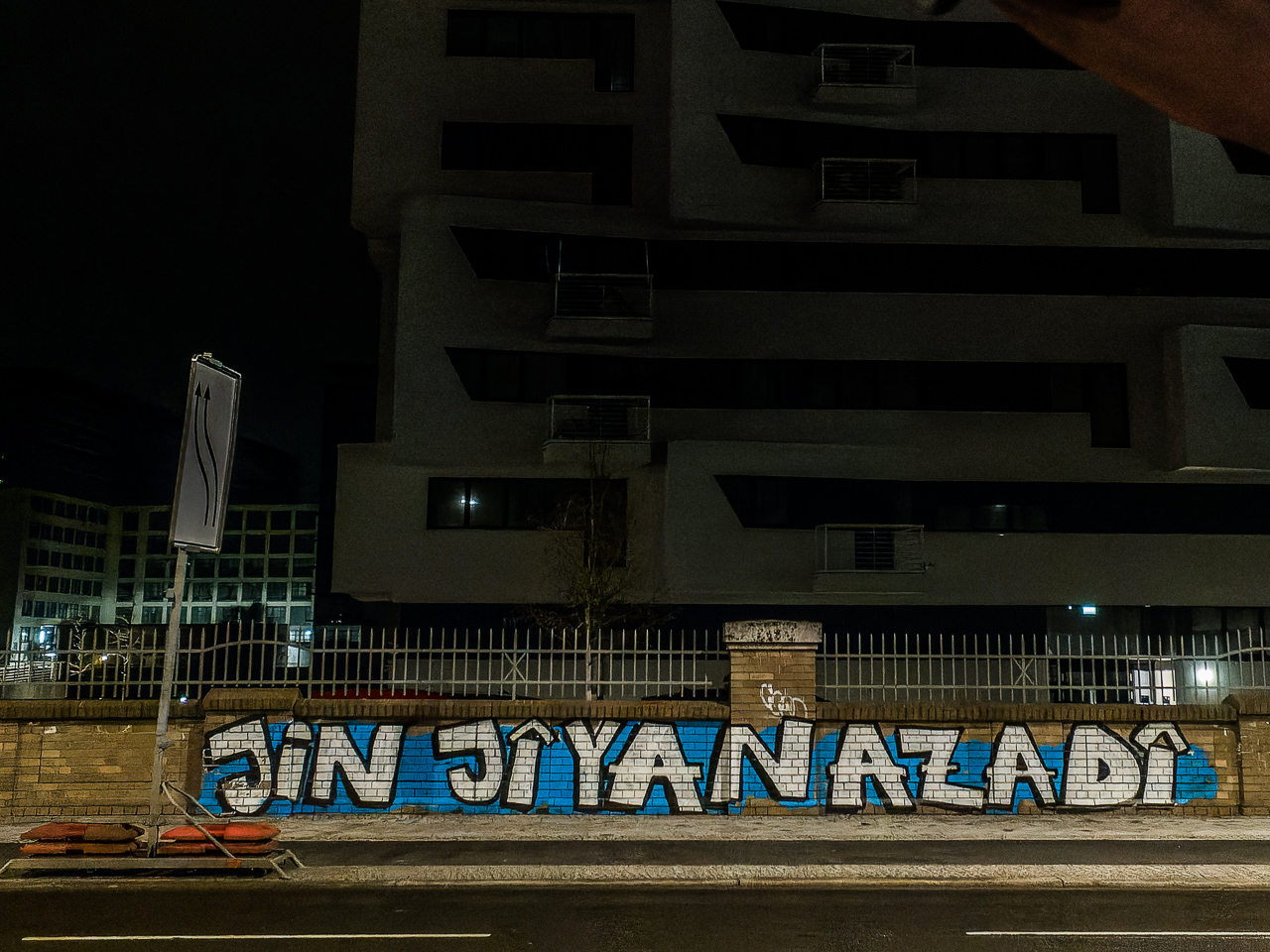Auf einer ZiegelMauer vor einem dunklen HochHaus ist auf blauen Grund weiß der iranische Schriftzug Jin, Jiyan, Azadî – Frauen Leben Freiheit gemalt