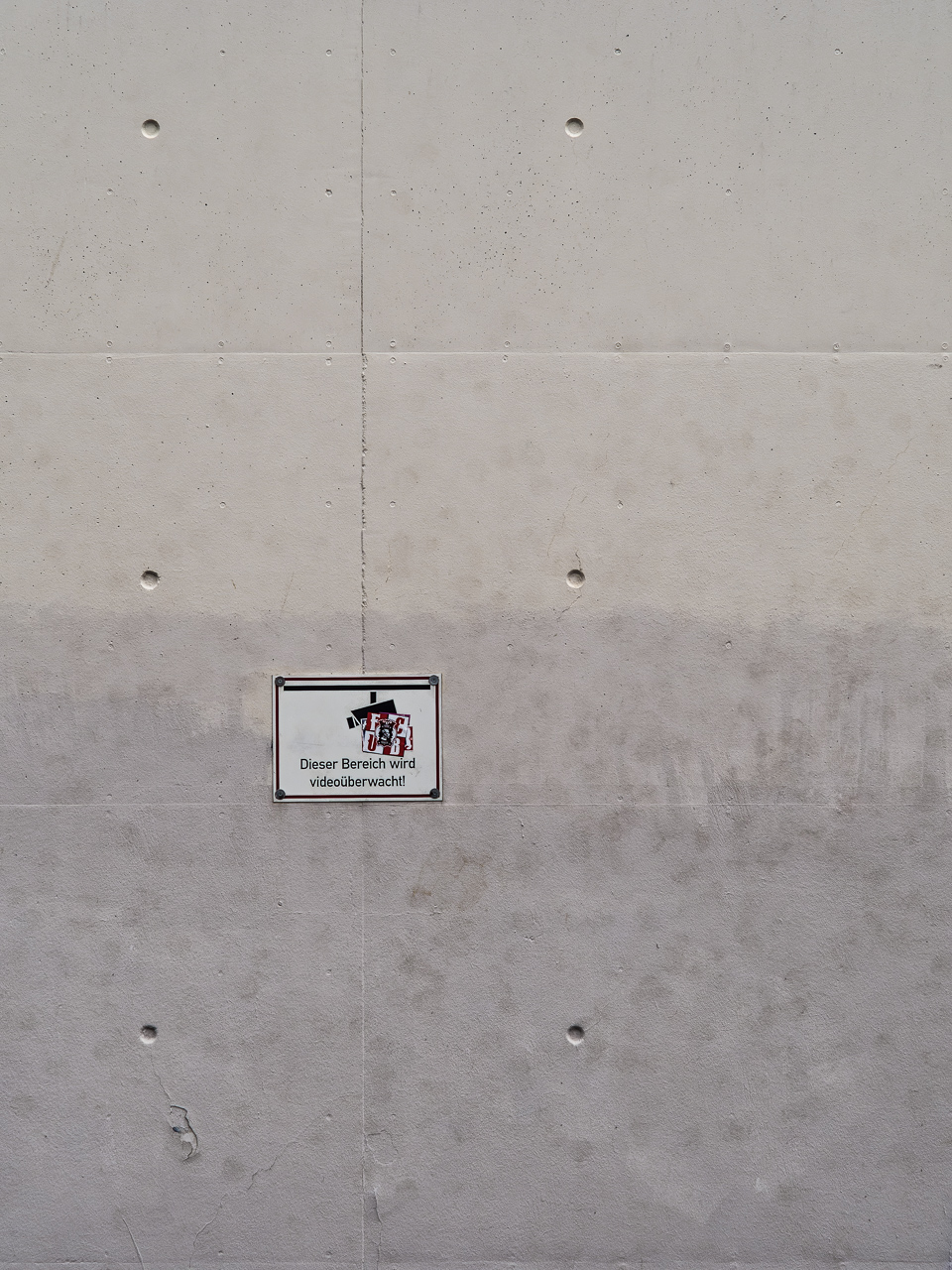 Weiß geputzte, etwas fleckige Wand, darauf ein Schild "Dieser Bereich wird videoüberwacht"