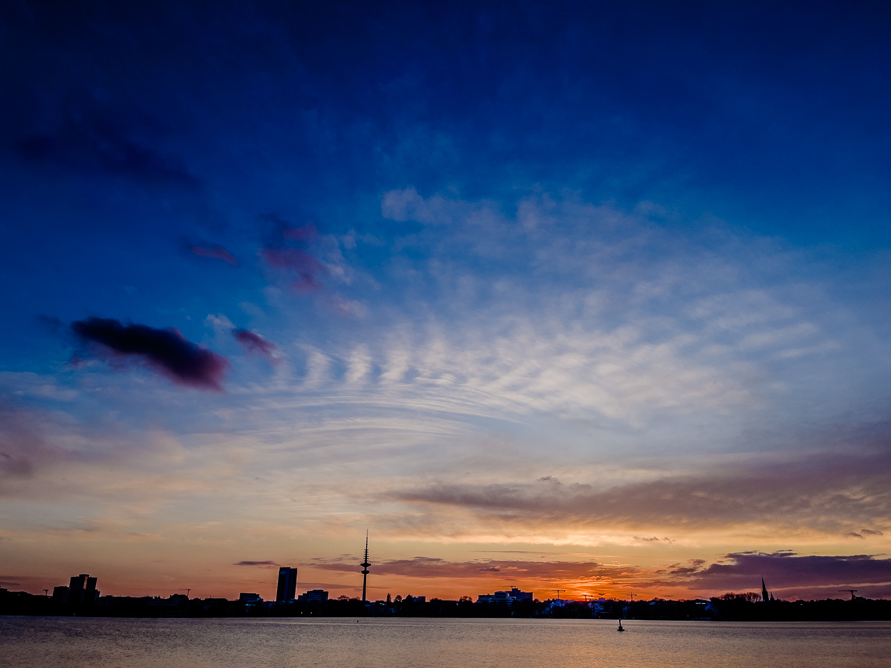 Städtischer See, SonnenUntergang. Silhoeutte von FernsehTurm und HochHaus, darüber vor dunkelBlauem Himmel ein gekrümmtes WolkenBand