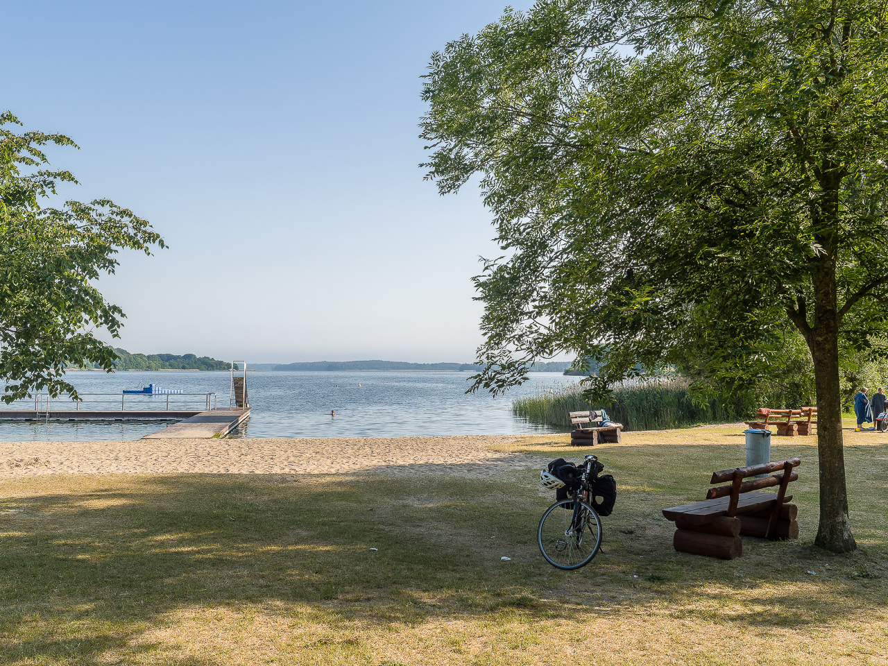 Ein StrandBad an einem See, Steg und Rutsche ins Wasser, im Vordergrund eine schattige SitzBank unter einem Baum