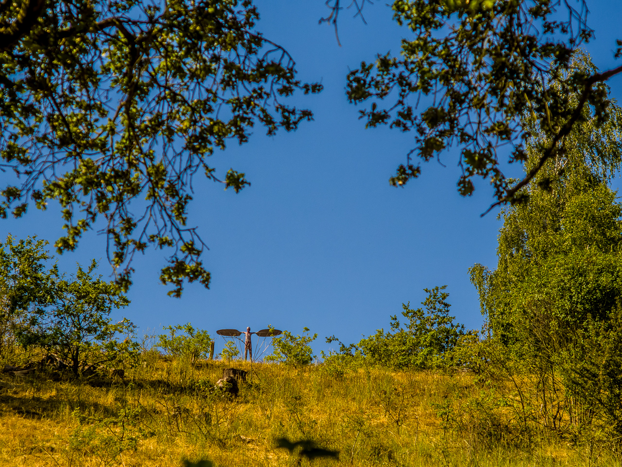 Ein mager bewachsener Hang von unten gegen blauen Himmel. Oben auf dem Berg steht eine Skulptur eines Menschen mit ausgebreiteten Armen, die wie Flügel aussehen.