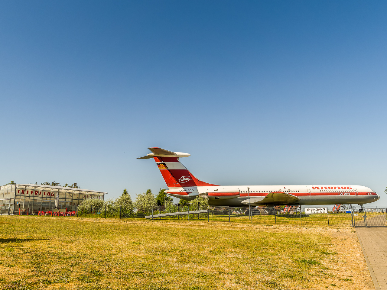 Eine PassagierFlugMaschine, rot und weiß, mit Kennung "Interflug" steht eingezäunt auf einer Wiese, daneben ein gläserner Pavillon mit Aufschrift "Interflug Café Airport"