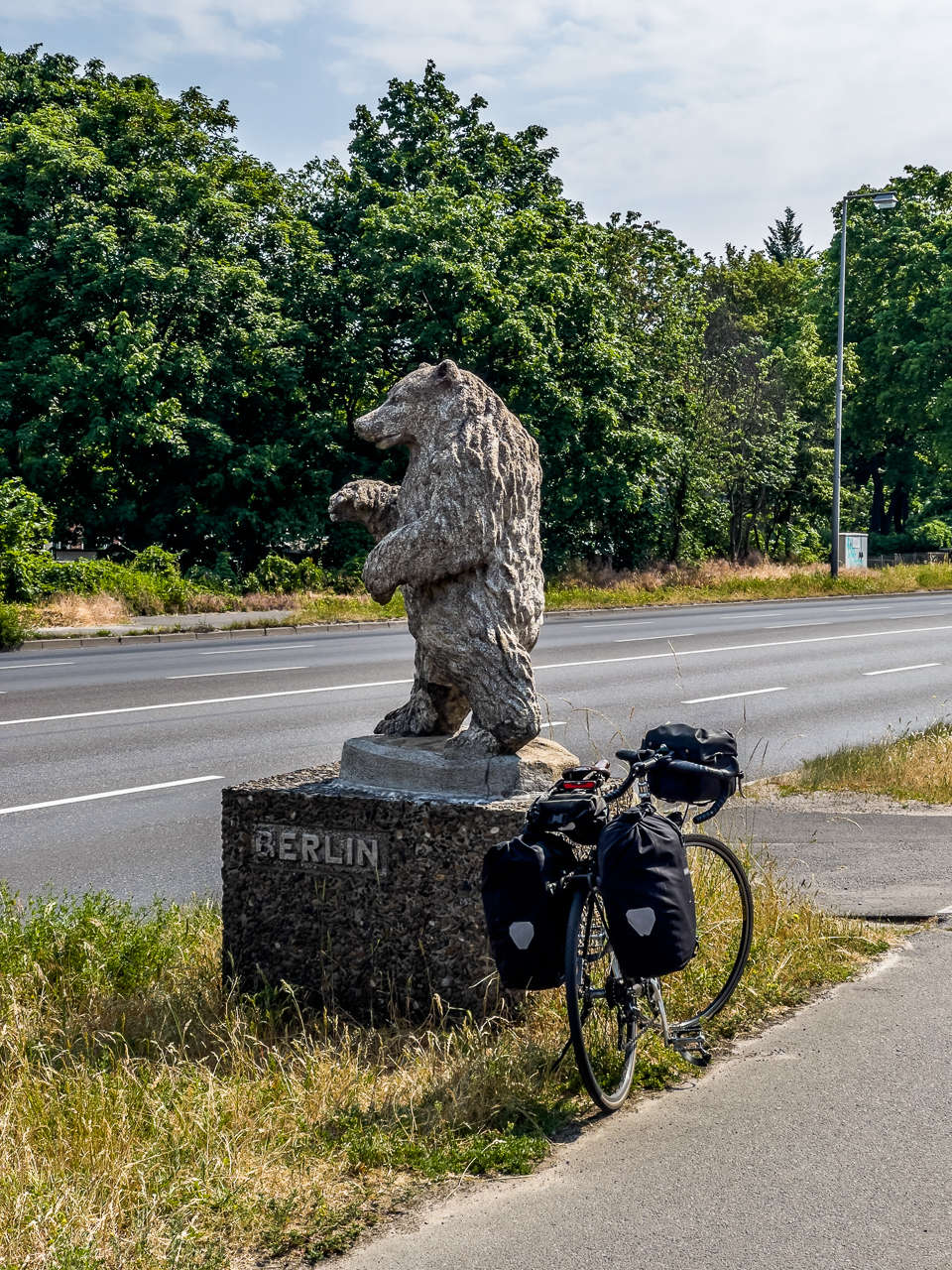 Skulptur eines Bären mit Beschriftung "Berlin" an einer breiten Straße, daneben steht ein Fahrrad mit ReisePackTaschen