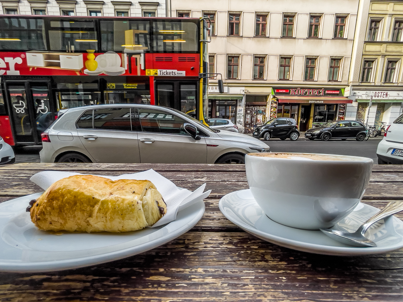 aus niedriger Perspektive fotografierte Teller mit einem SchokoCroissant und einer KaffeeTassee auf einer alten, abgeschabten HolzTischPlatte. Dahinter StraßenSzene einer Großstadt mit vielen Autos und einem DoppelStockBus