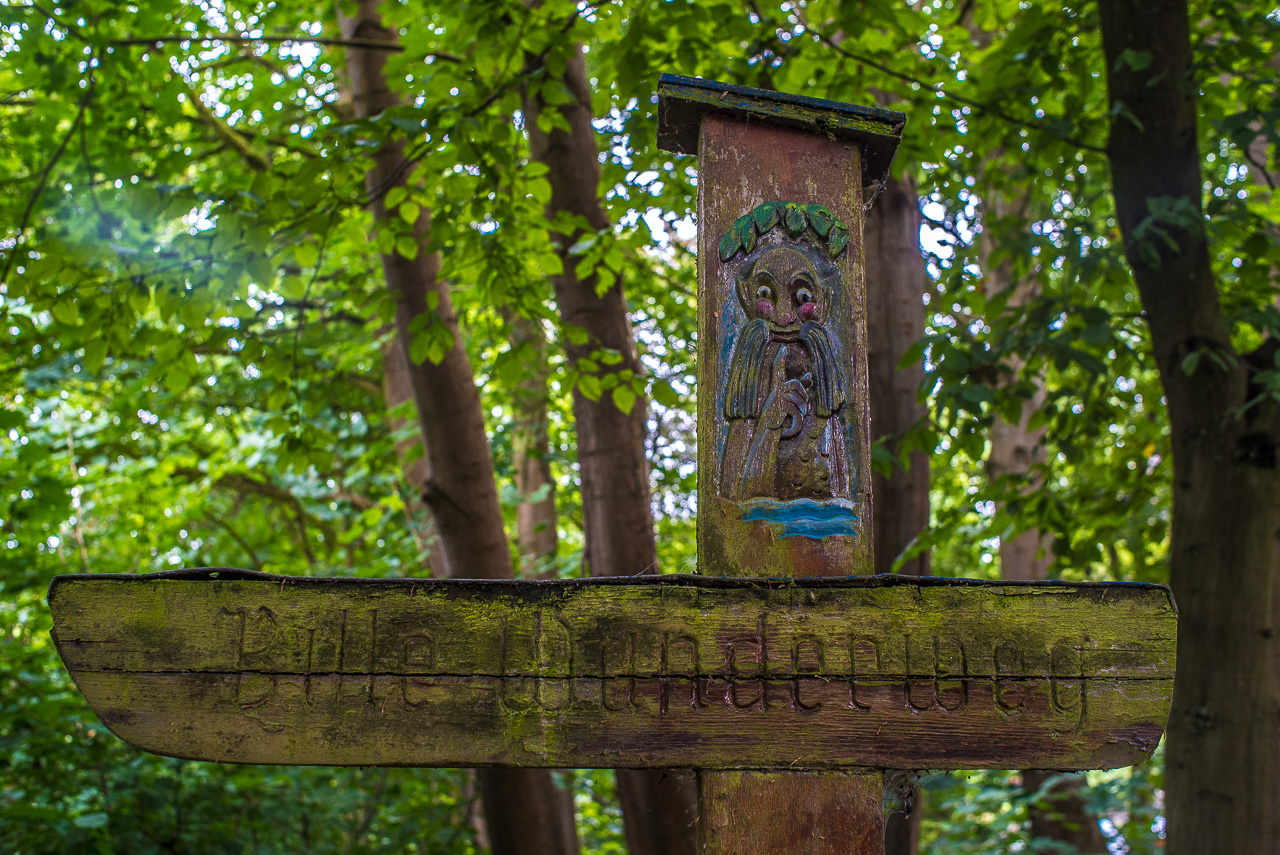 AltModischer Holz-WegWeiser in waldiger Umgebung, eingraviert "Bille-Wanderweg", darüber eine geschnitzte Figur mit roten Wangen und LorbeerKranz, die auf Wasser schaut.