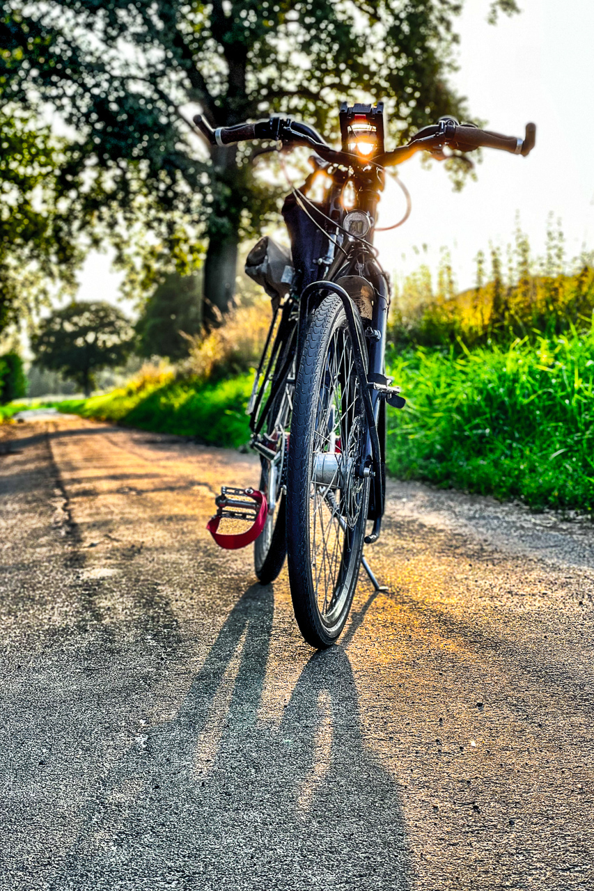 Ein schwarzes Fahrrad von vorn, auf einem AsphaltWeg am FeldRand, vor tief stehender Sonne.