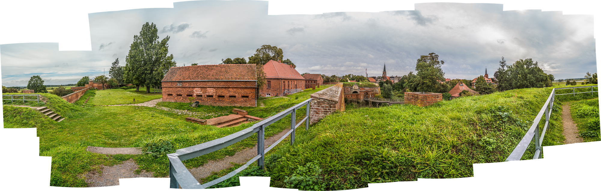 ZusammenGestückeltes Panorama einer FestungsAnlage mit gras-bewachsenen Wällen, BacksteinGebäude, im Hintergrund eine alte Stadt mit mehreren spitzen KirchTürmen