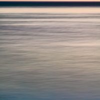 MeeresSpiegel in warmen AbendLicht. Das Foto ist ein enges HochFormat, der unscharfe Horizont liegt etwa an der oberen ViertelTrennung. Darunter weiche, durch sehr lange Belichtung verwischte Wellen, die in der Farbe von dunklem GrünBlau vorn zu pastelligem Gelborange verlaufen. Der Himmel ist ein etwas intensiveres Orange.