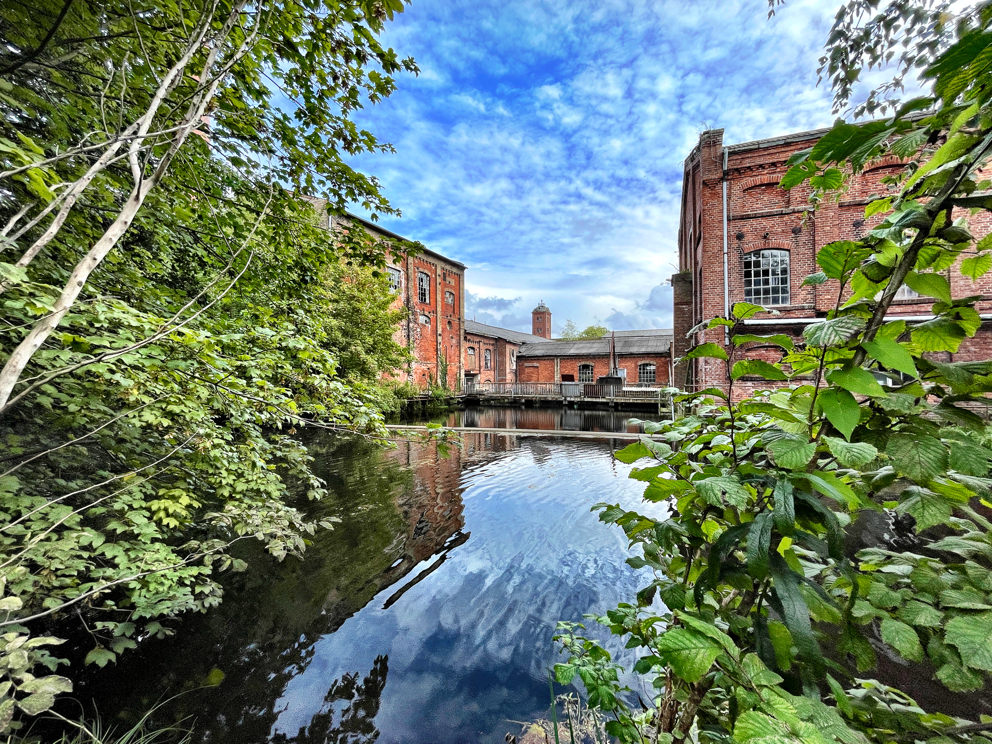 Backstein-FabrikAnlage an einem Wasserlauf, umgeben von vielen Bäumen. SchäfchenWolken an blauem Himmel.
